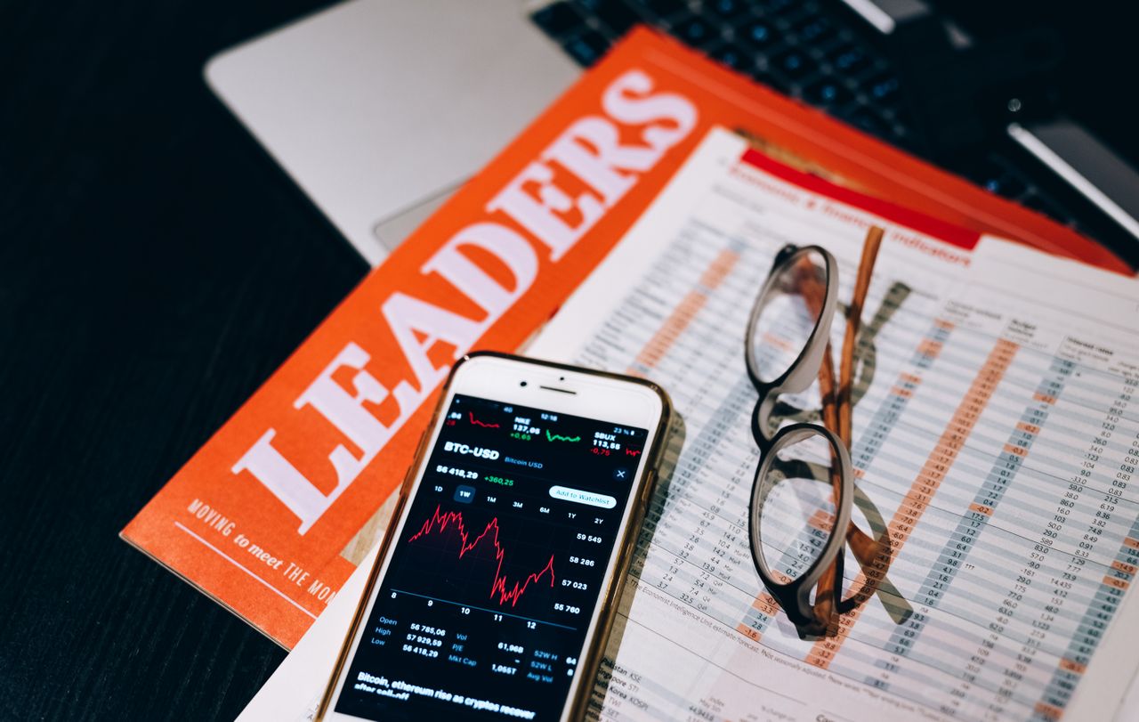 A close-up of a smartphone with cryptocurrency data next to financial reports and a magazine titled 'LEADERS'.